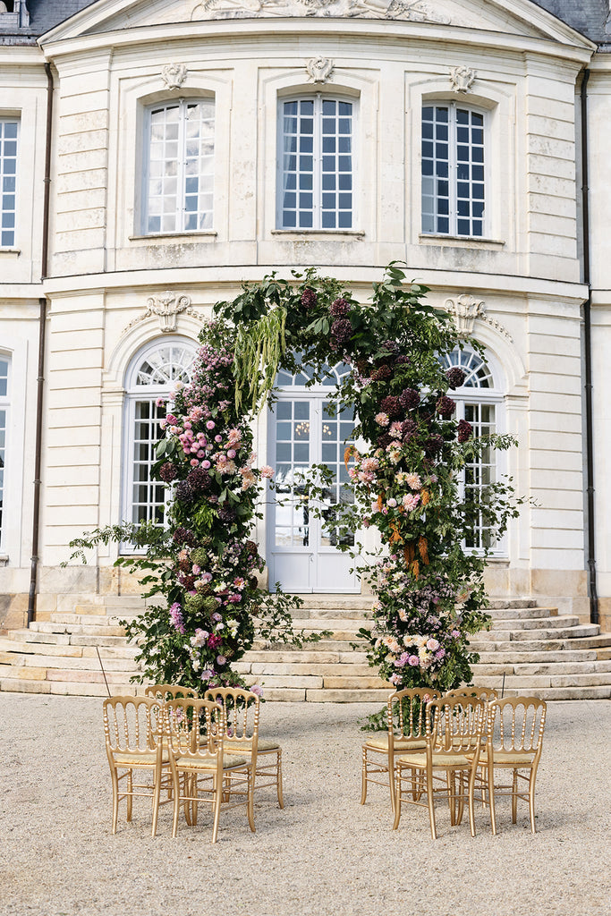 A floral archway outside of a European estate with gold chairs.