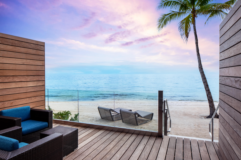 The oceanfront view of the Casa Marina Key West, Curio Collection by Hilton. A patio and lounge chairs look out onto a beach with a purple-blue sunset.