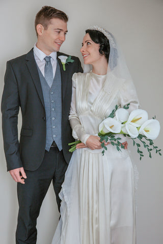 1920s looking couple gazing lovingly at each other with the bride holding a bouquet of cascading paper calla lillies and wearing an intricate paper lily flower crown with her white veil sweeping down from her black wavy hair to her white satin dress