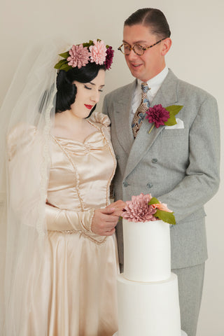 1940s looking bride and groom. Bride is wearing a peachy apricot satin dress with a purple paper dahlia flower crown over her veil. The groom is wearing a grey breasted suit with burgundy paper dahlia button hole. They are holding hands in front of a cake