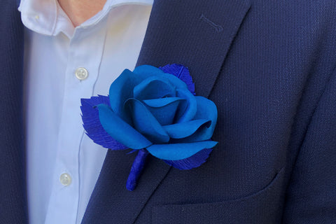 Bright blue rose boutonniere with darker bright blue leaves attached to a navy jacket lapel, against a white collared mens shirt. 