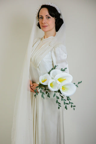 1920s bride with long cascading white veil with tiny paper lily crown, long satin dress, holding a sheath bouquet of white crepe paper calla lilies
