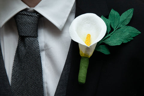 Single white calla lily paper flower button hole with forest green leaf vine attached to the lapel of a black dress jacket, over the top of a white dress shirt with coordinating grey tie and vest