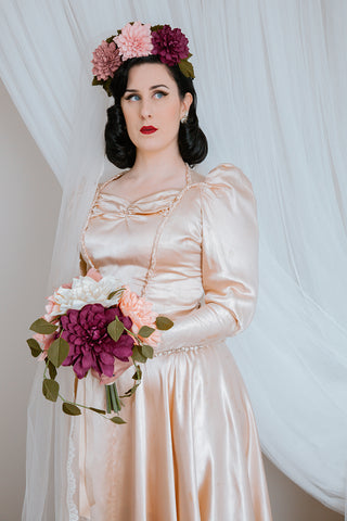 1940s styled bride with a crown of paper dahlias over the top of a vintage long veil, light peach satin wedding dress and hoping a volumous bouquet of paper dahlias in cream, mulberry and peach 