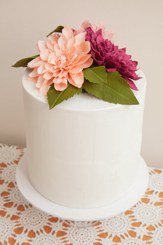 Salmon pink and burgundy crepe paper dahlias with olive green crepe paper dahlia leaves perched on top of a tall white single tiered fondant wedding cake