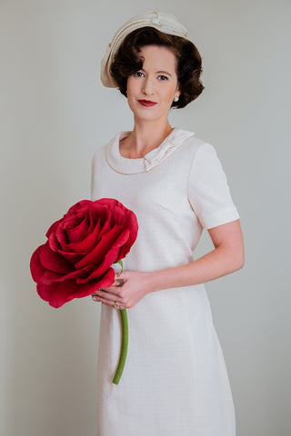 1960s bride in cream slouched hat, cream short sleeved vintage dress, holding a giant crepe paper red rose