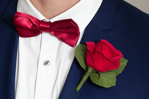 Single red rose crepe paper boutonniere with three olive green leaves, pinned to navy blue jacket lapel, that sits over the top of a white dress shirt with matching dark red bow tie