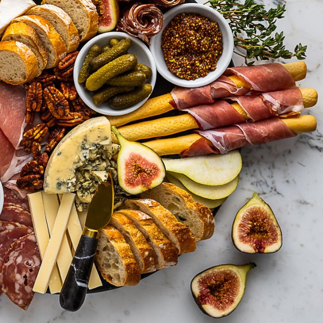 Black cheese board and marble cheese knife with fresh figs, meat, and cheeses.