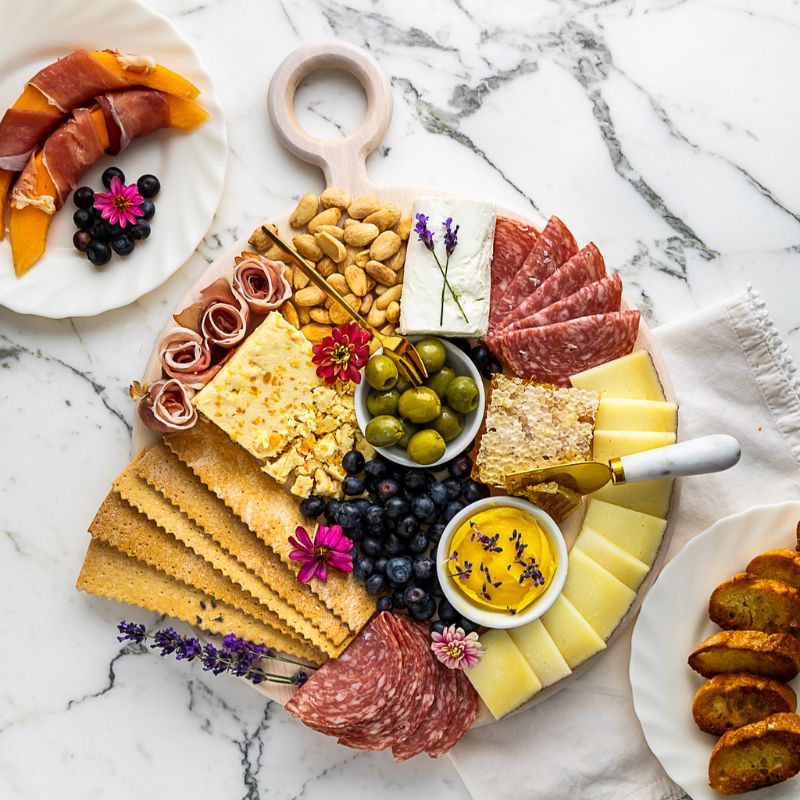 Round white serving board with cheese, charcuterie, and summery accompaniments.