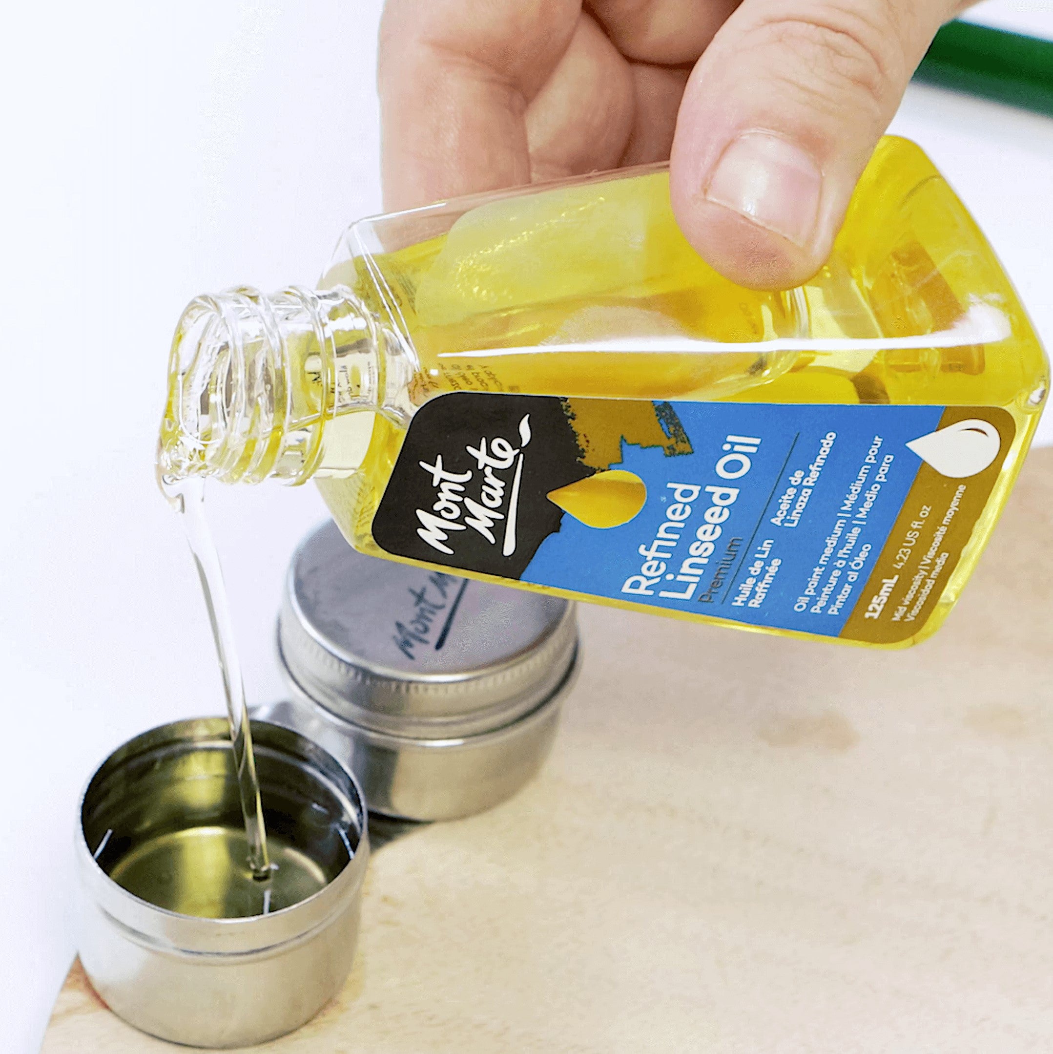 A hand pouring Mont Marte Refined Linseed Oil into a metal lid on a wooden board.