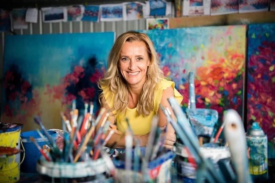 Artist Belinda Nadwie in a bright yellow blouse smilling in her studio in front of paint brushes and paint buckets with her artworks behind her.