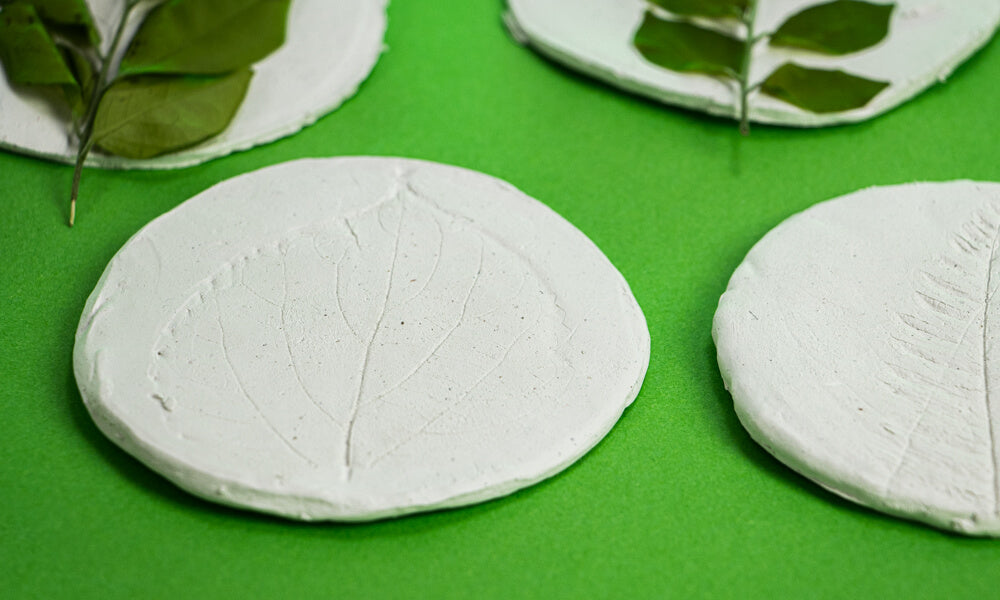Four circles of air dry clay with a leaf imprint printed on top on a green background.