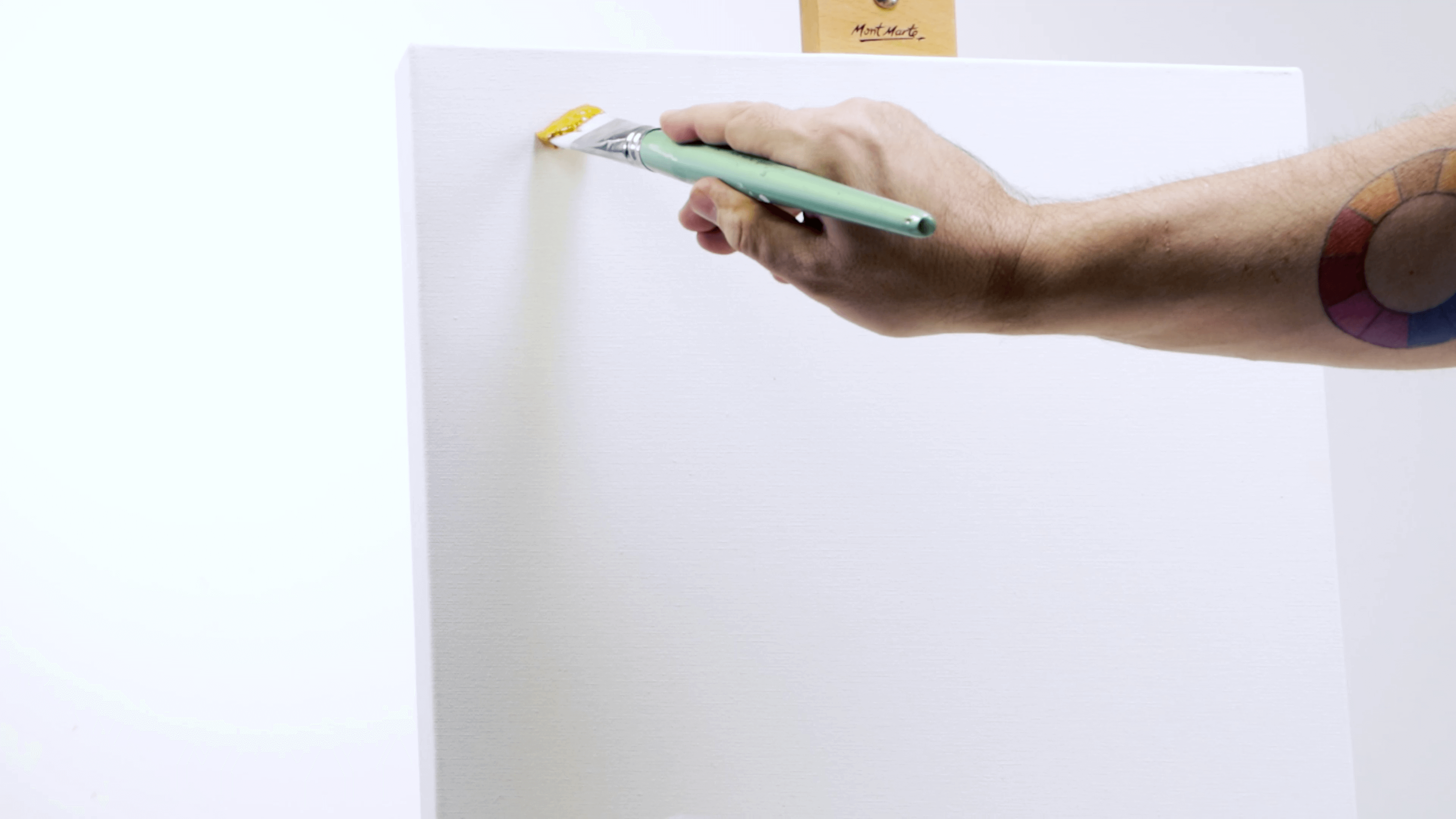 Hand holding a filbert paint brush applying yellow paint to a canvas on a wooden easel.