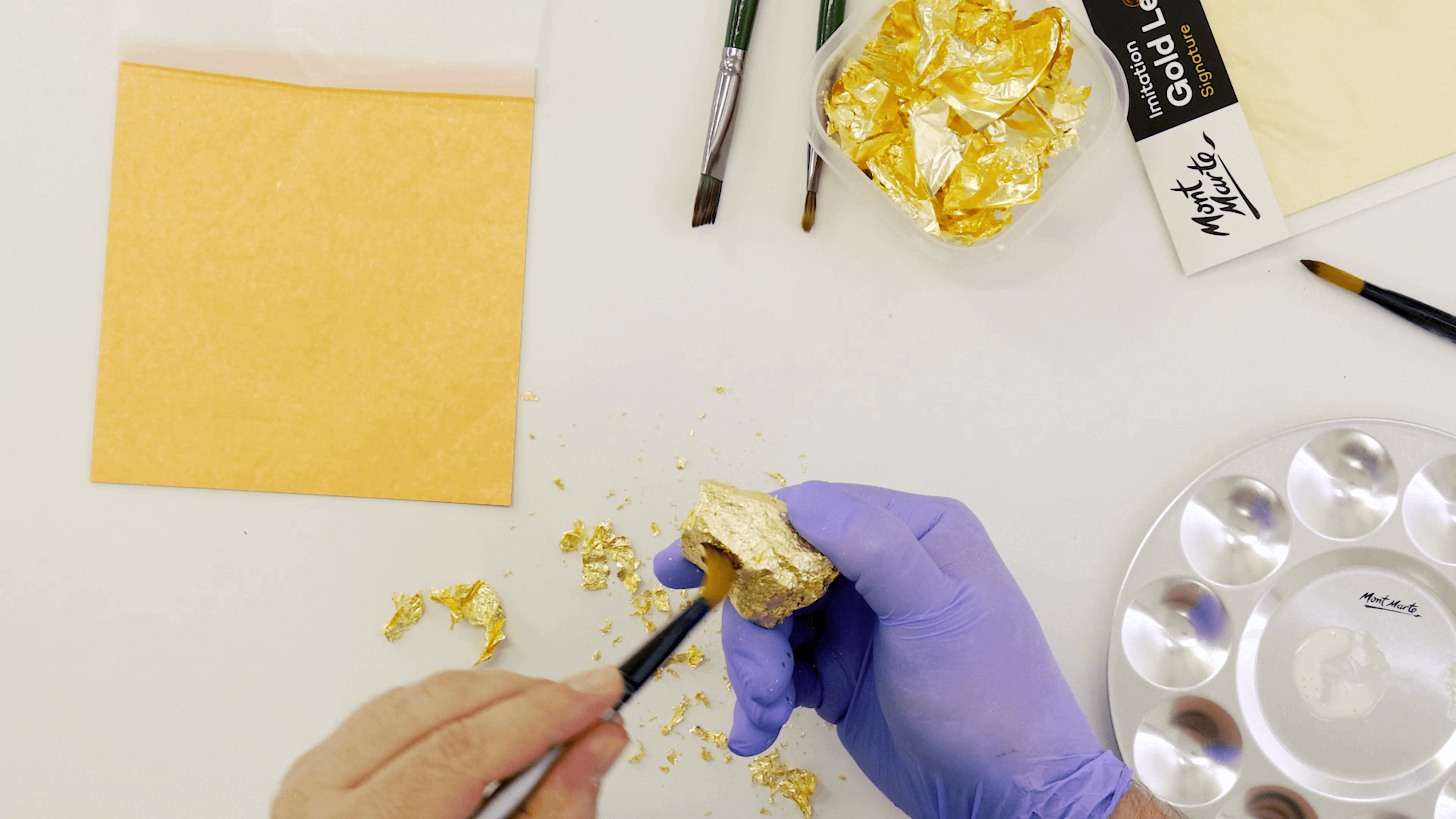 Person brushing gold leaf on a rock wearing a purple glove, in a busy workspace.