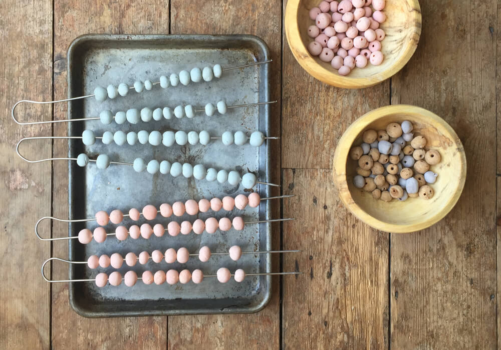 A baking tray with polymer clay beads laying on wire skewers on top with loose beads next to the tray.