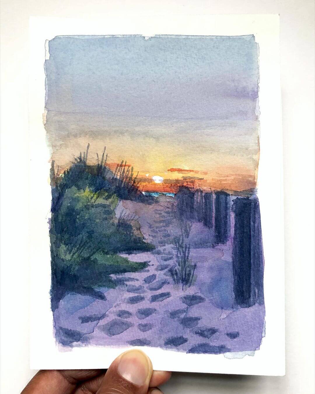 A hand holding a sketch book with a completed beach at sunset painted in watercolours.