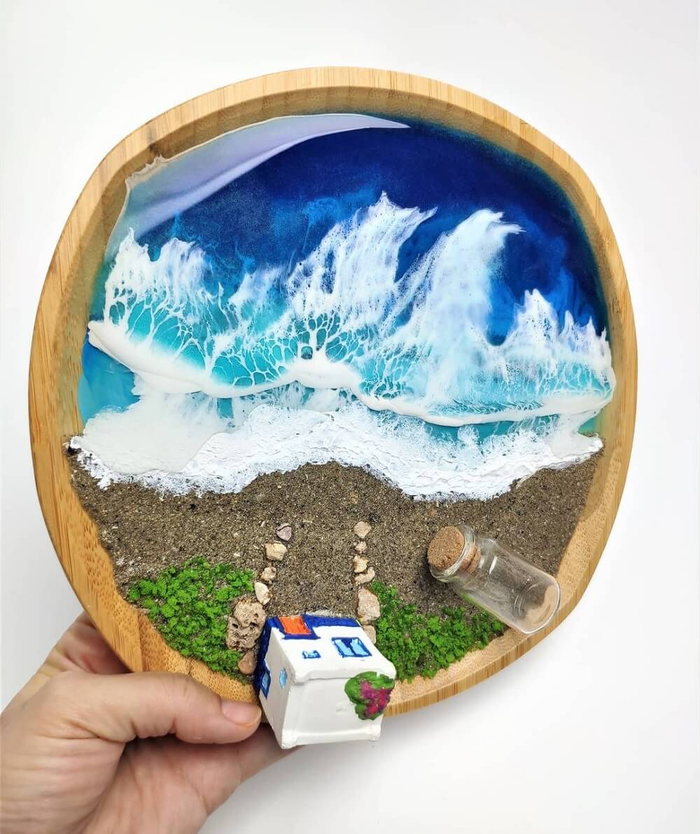 Hand holding a diorama of a beach built in a wooden circle with found objects glued to it.