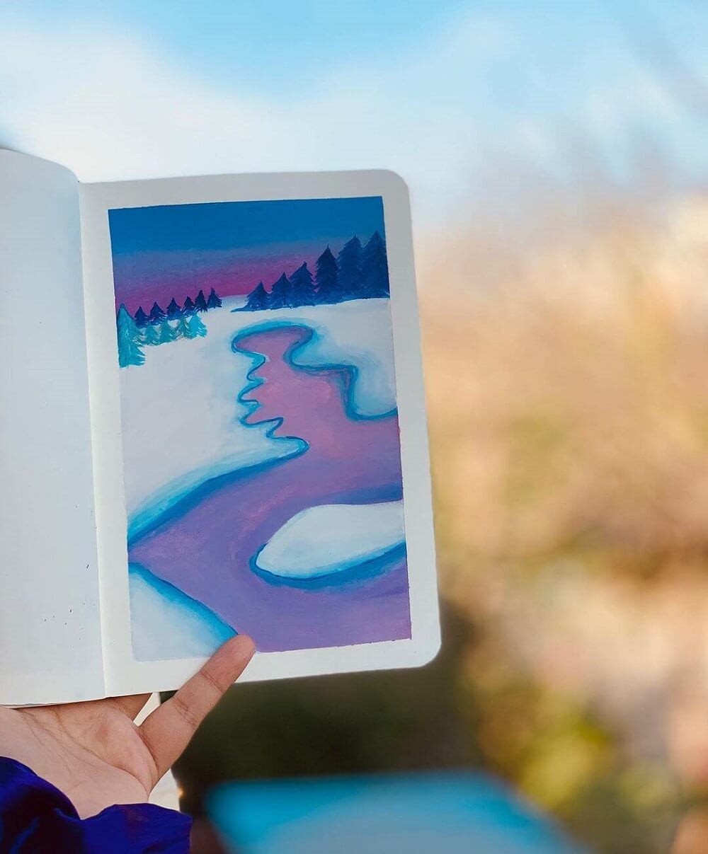 Hand holding a sketchbook of a gouache artwork with a icy lake.