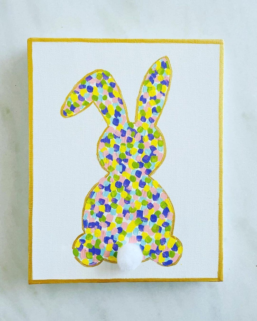 A bright bunny artwork painted on canvas with yellow, blue and purple colours and a gold frame with a cotton bud tail.