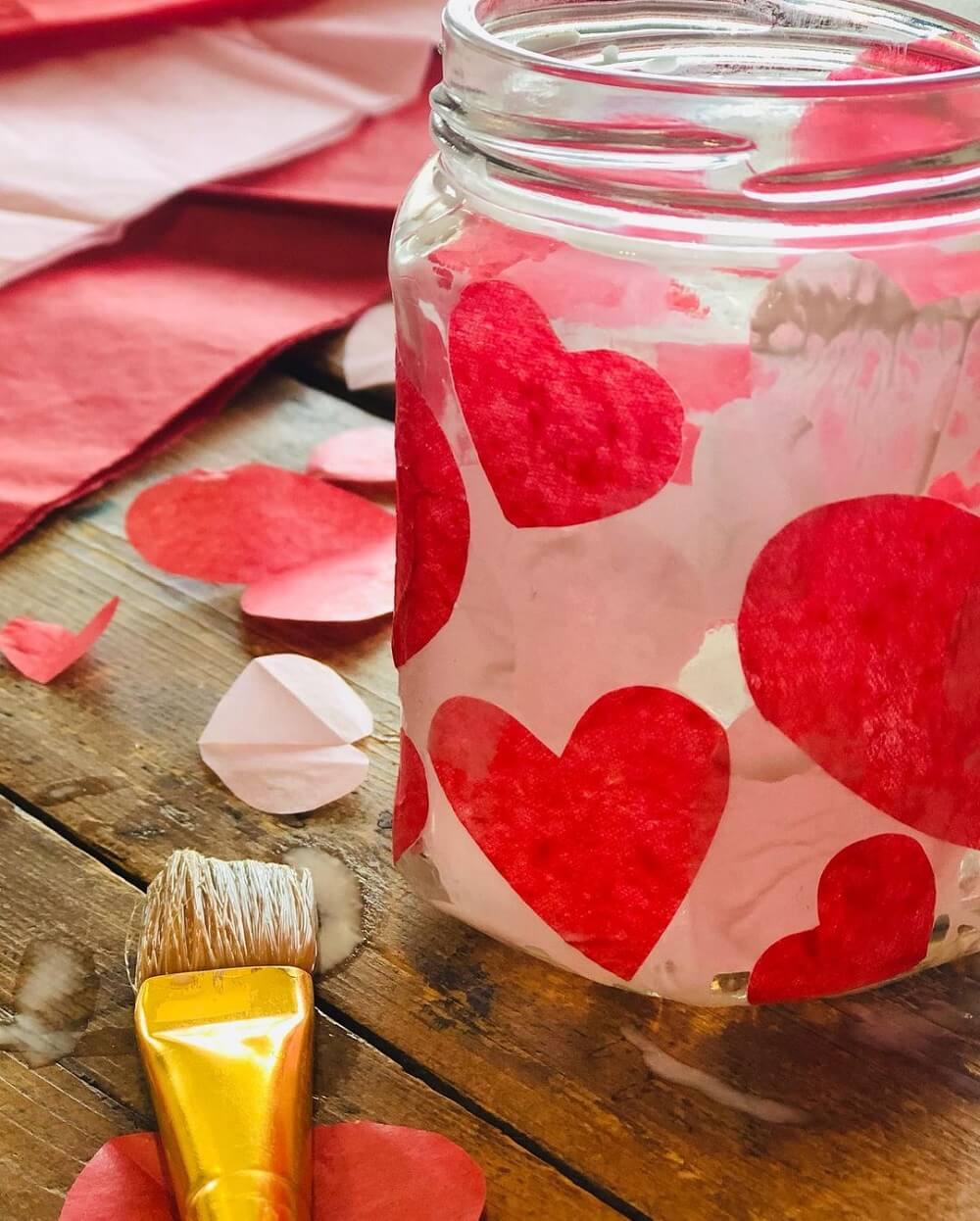 Mason jar with hearts glued on next to red tissue paper and a gold taklon brush.