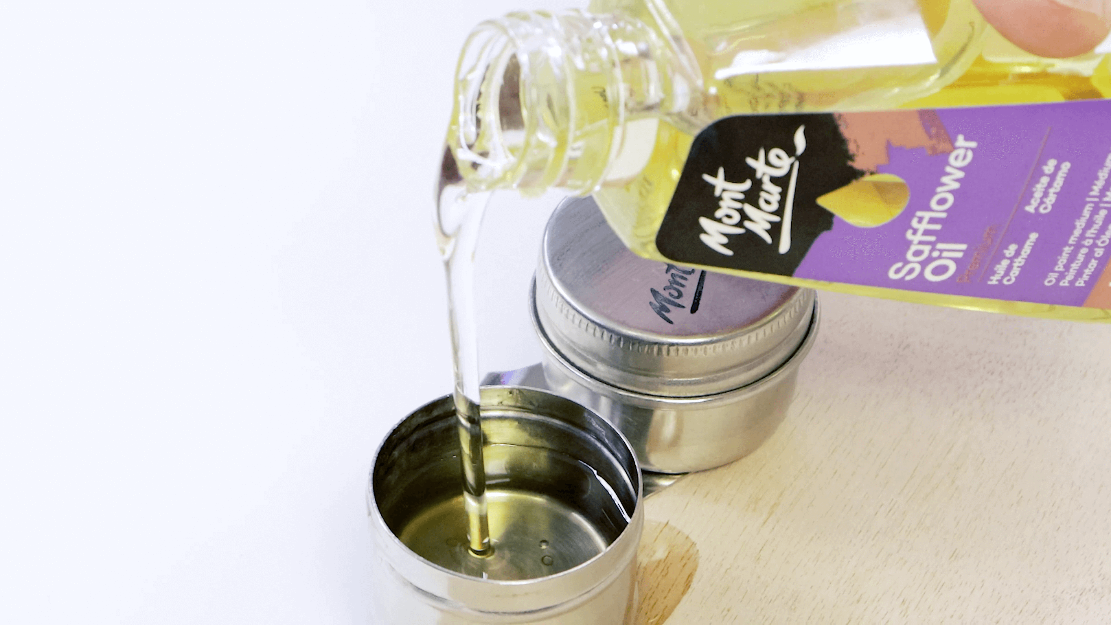 A hand pouring Mont Marte Safflower oil into a metal lid on a wooden board.