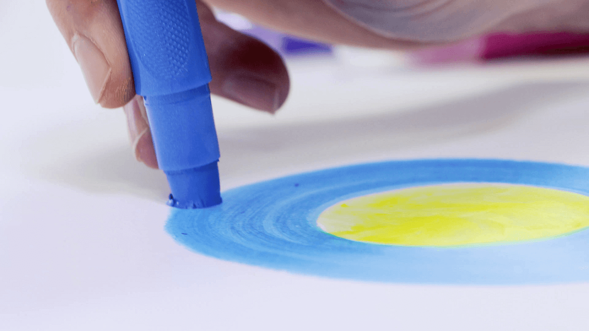 Hand holding a blue Mont Marte twistable metallic gel stick and colouring in around a yellow circle.