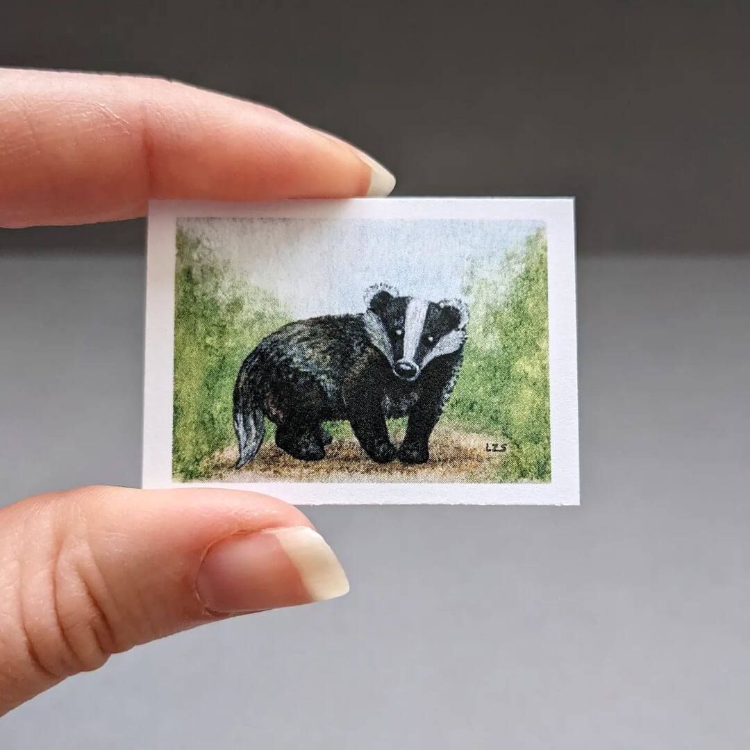 Hand holding a very small, realistic watercolour artwork of a badger.