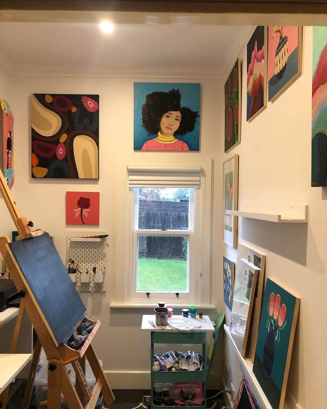 Colourful artwork hanging on walls above a small window and a blue trolley with art supplies on the side.