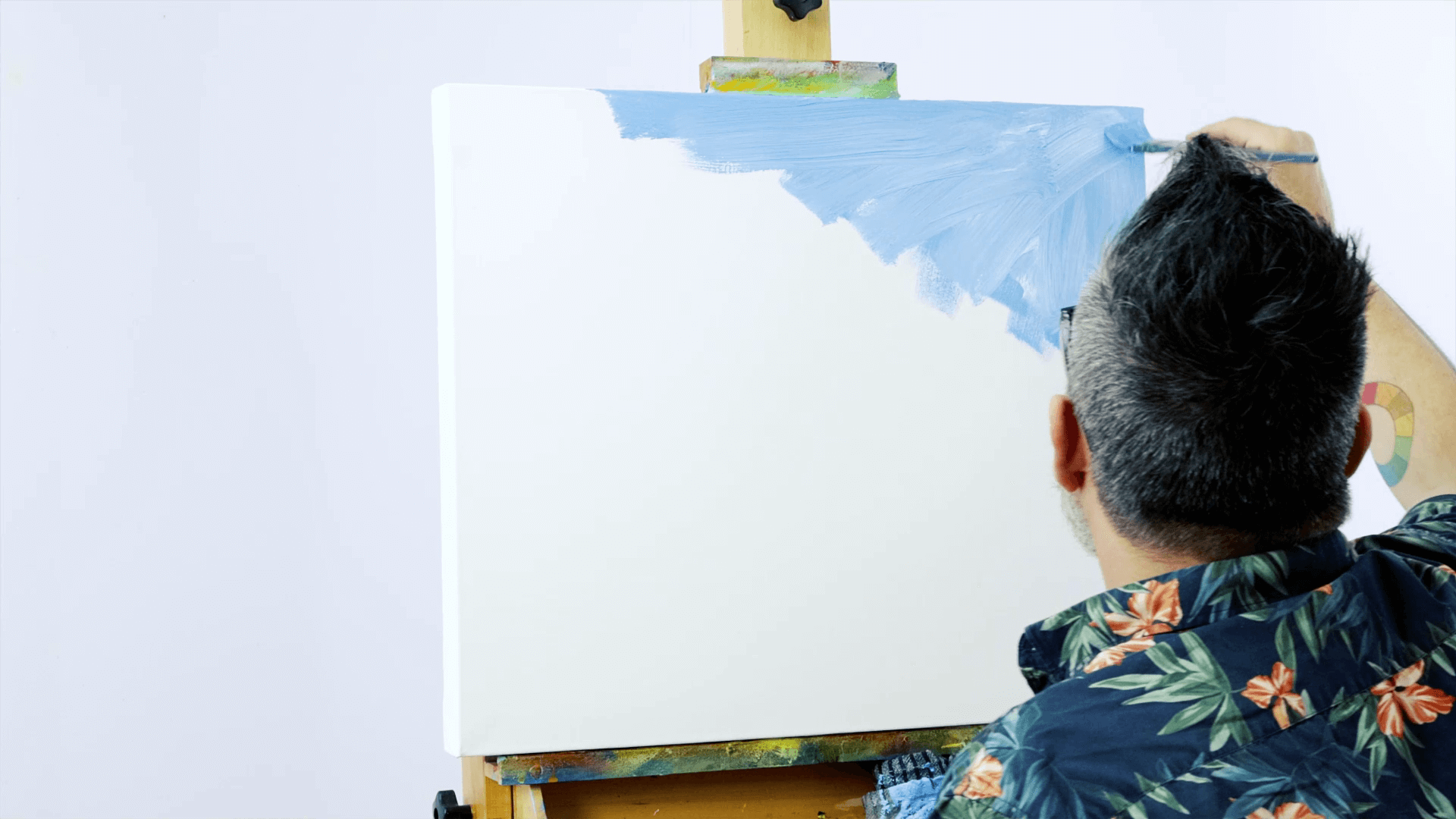 Man paints a white canvas on a wooden easel with blue acrylic paint, beginning from the top corner.