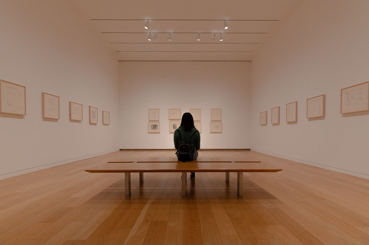 Person sitting on a wooden bench in an art gallery looking at the artworks on the walls.