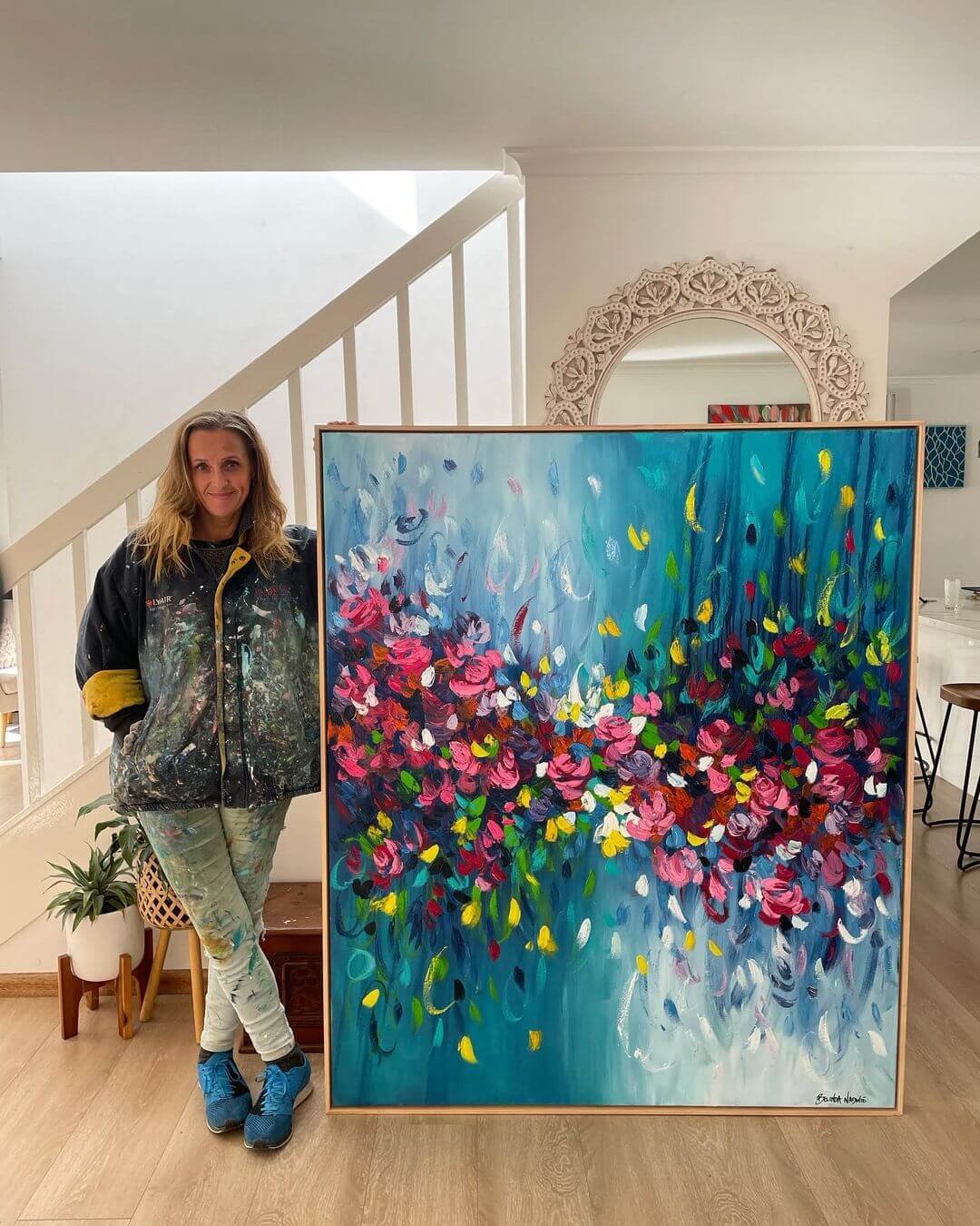 Belinda holding a large artwork of flowers in her home. Wearing jumper and jeans with paint splats in a modern home with white walls.