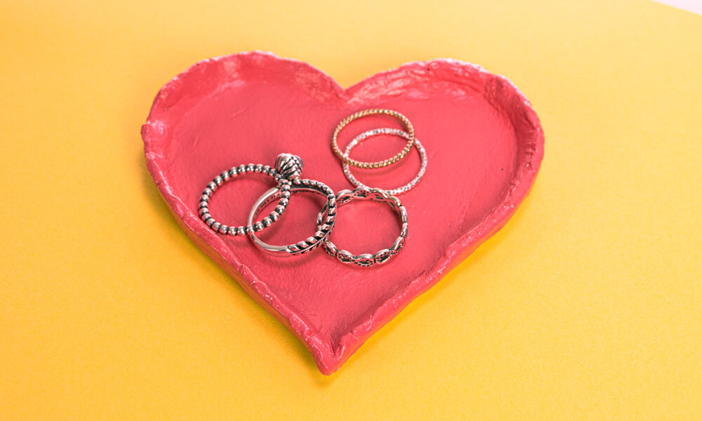 A love heart shaped air dry clay trinket dish painted red, with rings in the dish, on a yellow background.