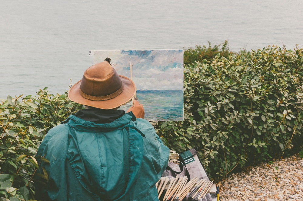 Person wearing a hat and green rain coat painting near a lake.