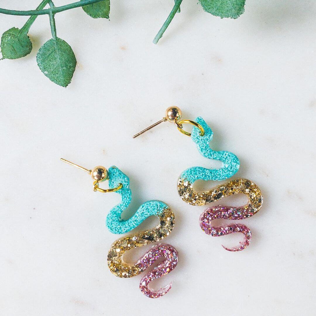 Two gold snake earrings with teal, copper and pink colours on them.