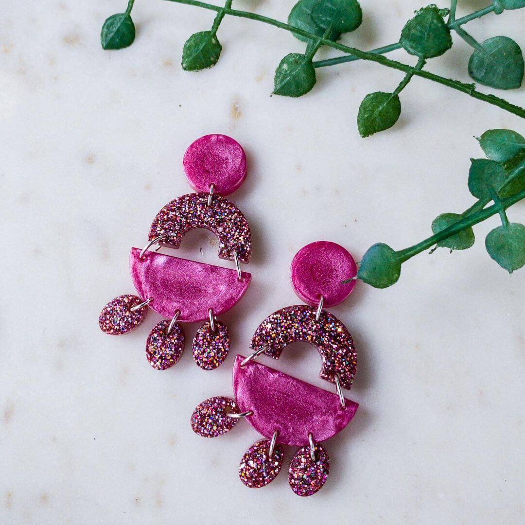 Pink glittery earrings made from resin with tear drop shapes next to a green plant.