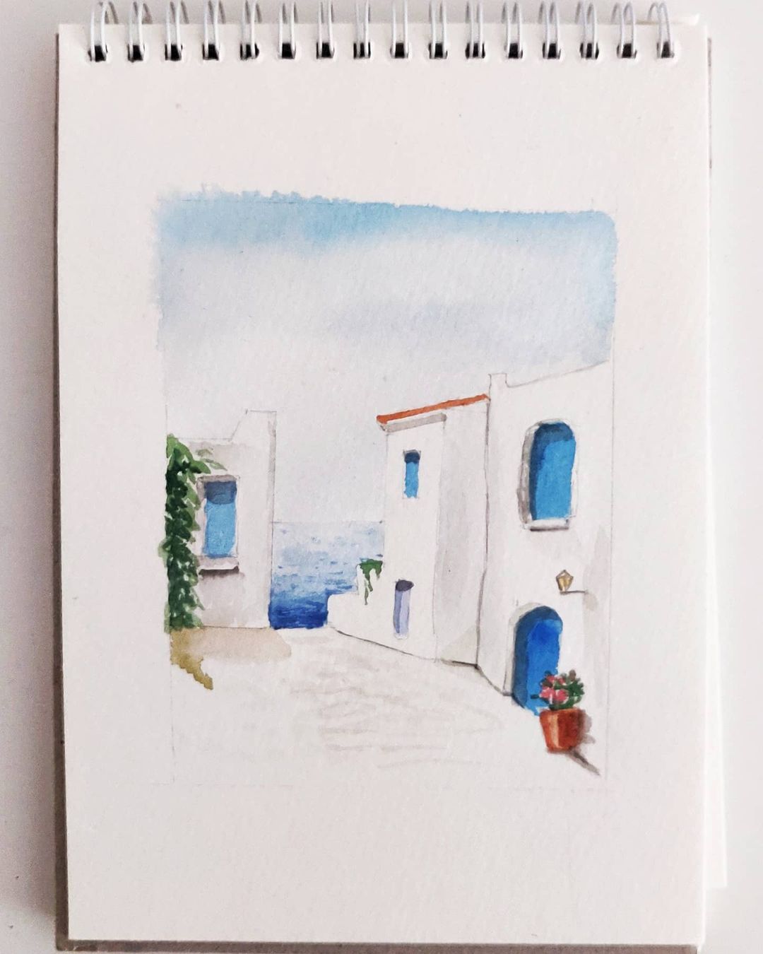 Santorini watercolour of a white building with blue windows and doors near the Mediterranean ocean.