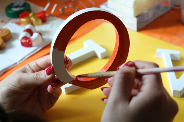 Person with red nails, painting a wooden letter with orange paint.