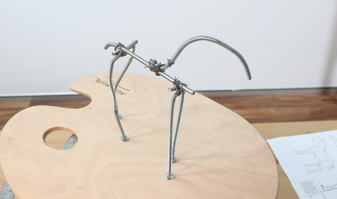 A wire armature of a horse is screwed onto a wooden palette.