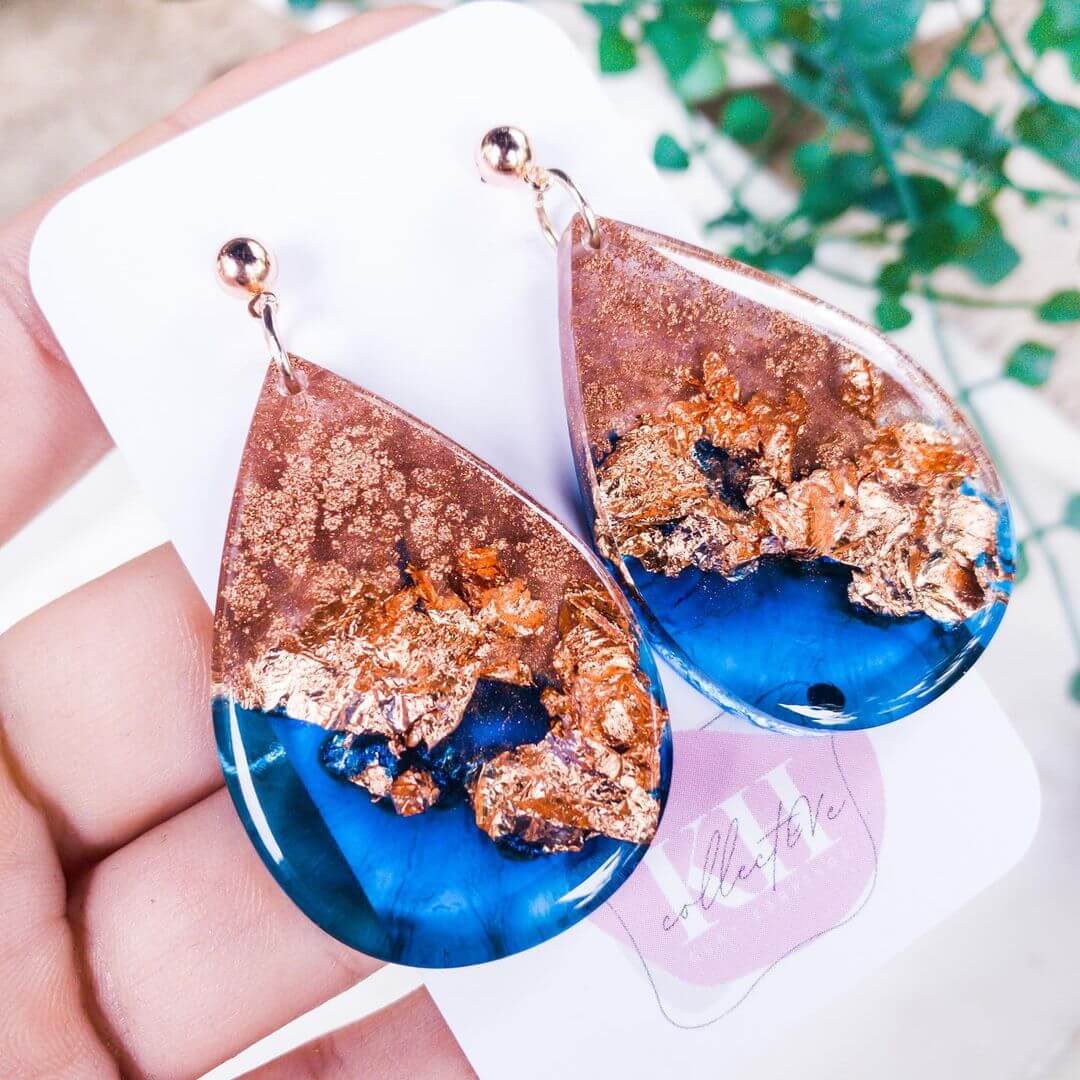 Hand holding blue and copper earrings made from resin near a green plant.