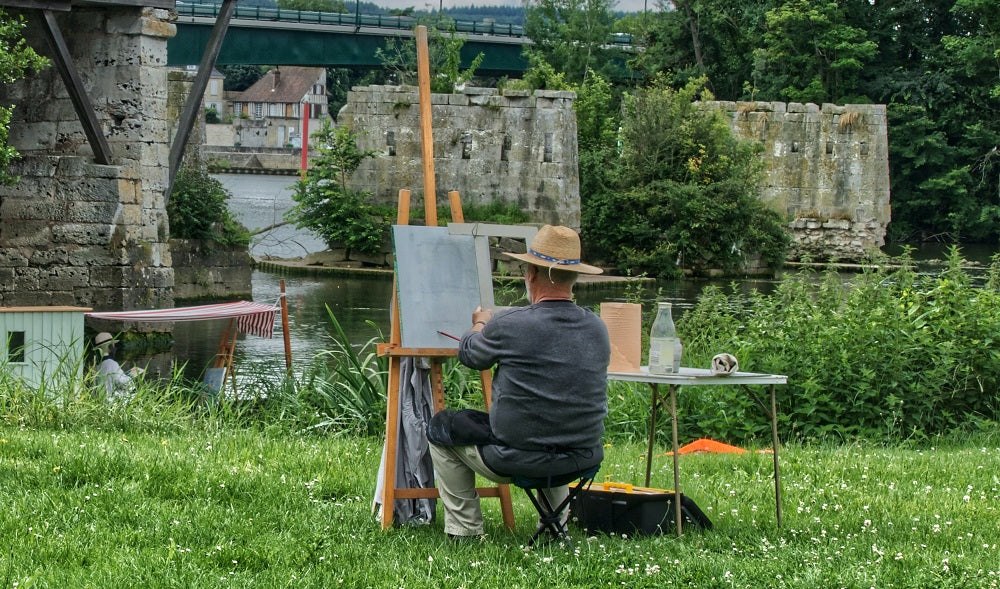 Man in a straw hat sits on a stool in a green field and paints a bridge.