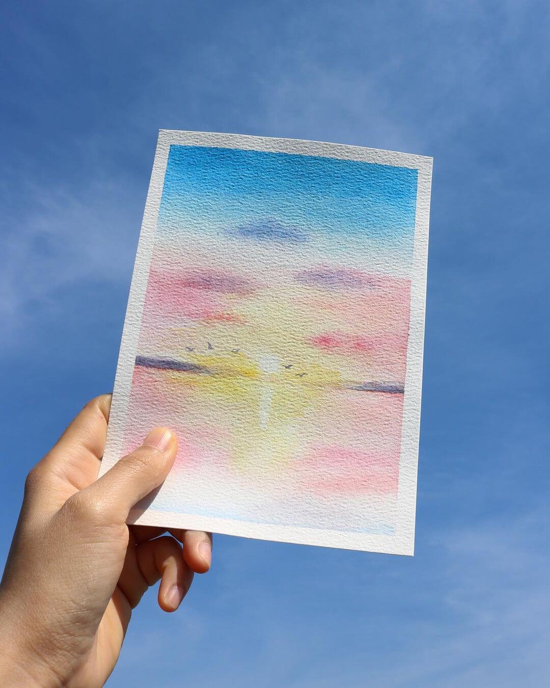 Hand holding up a sunset watercolour painting in the blue sky.