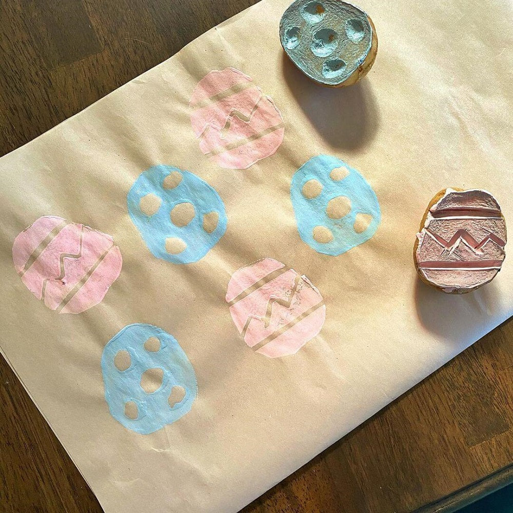 A potato sliced in half with different designs carved into each potato with pink and blue paint on each that has been stamped onto paper.