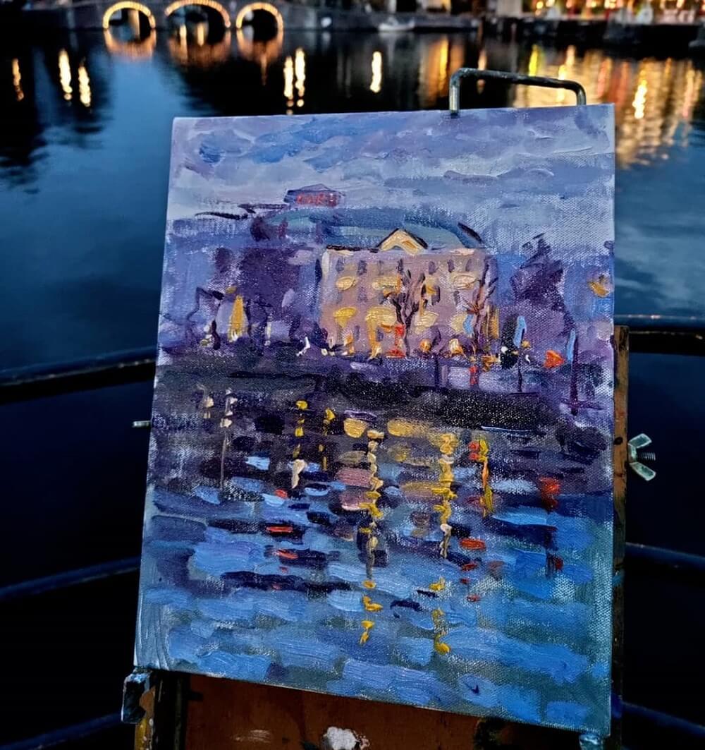 An evening oil painting of Amsterdam with lights reflecting off the river.