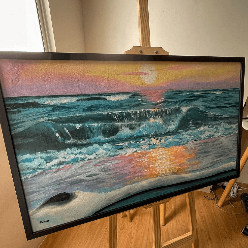 A realistic ocean at sunset, of pinks, blues and yellows painted on acrylic standing on an easel.