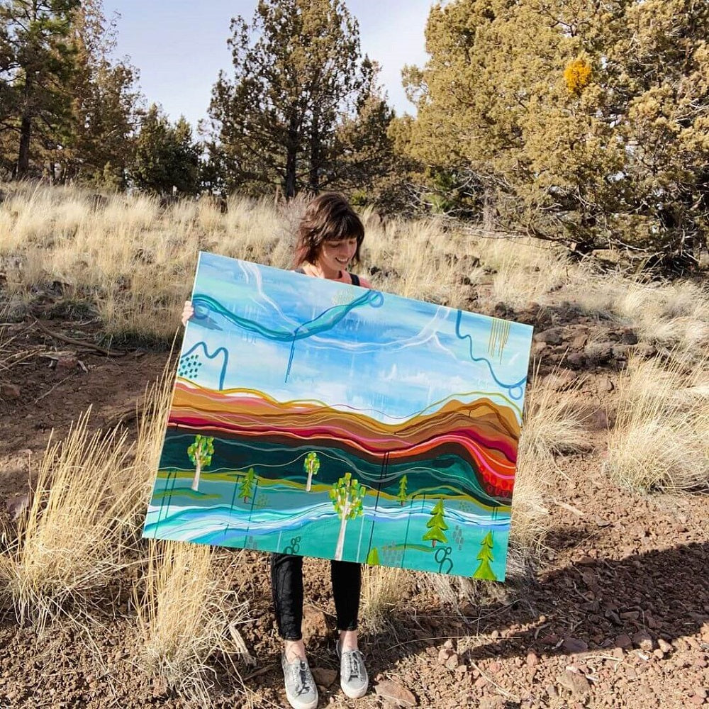 Artist Kristine Cooper standing in a large field, holding a colourful, large abstract painting of a landscape.