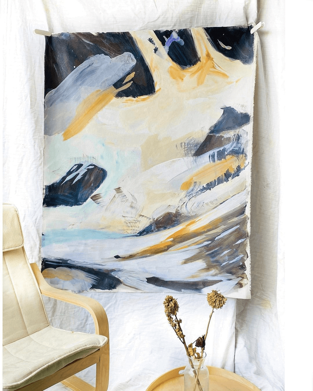 An abstract linen painting of navy, gold and white paint next to a vase of native flowers and a chair.