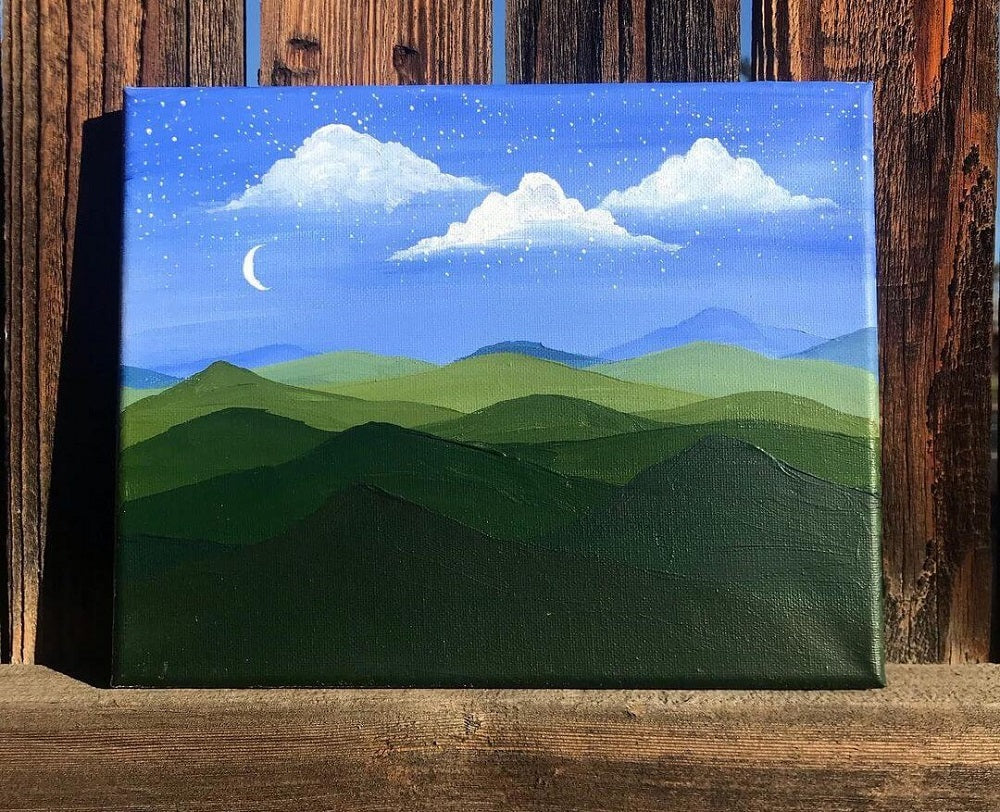 A painted canvas of a blue sky with small stars and green mountains underneath, canvas sits on a wooden board.