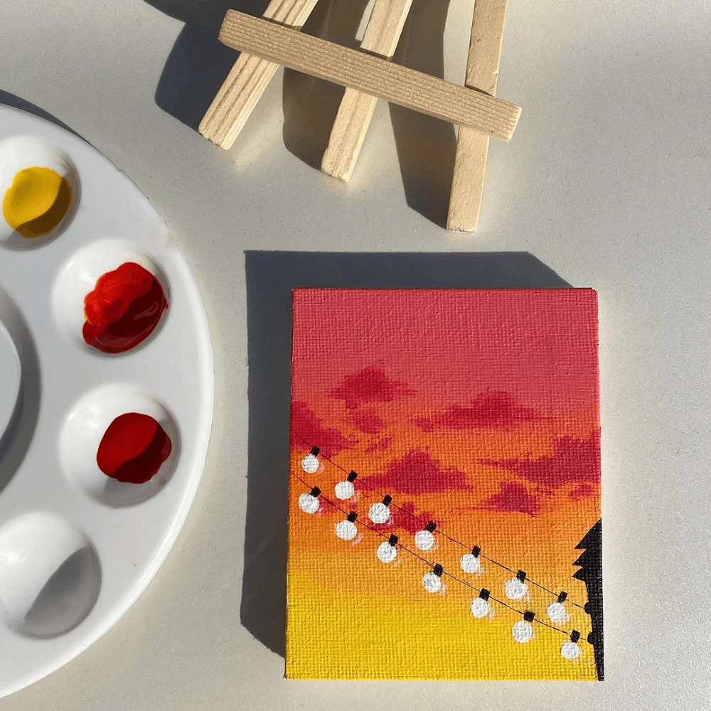 A palette of red and yellow paint next to a small canvas of a sunset landscape of red and white.