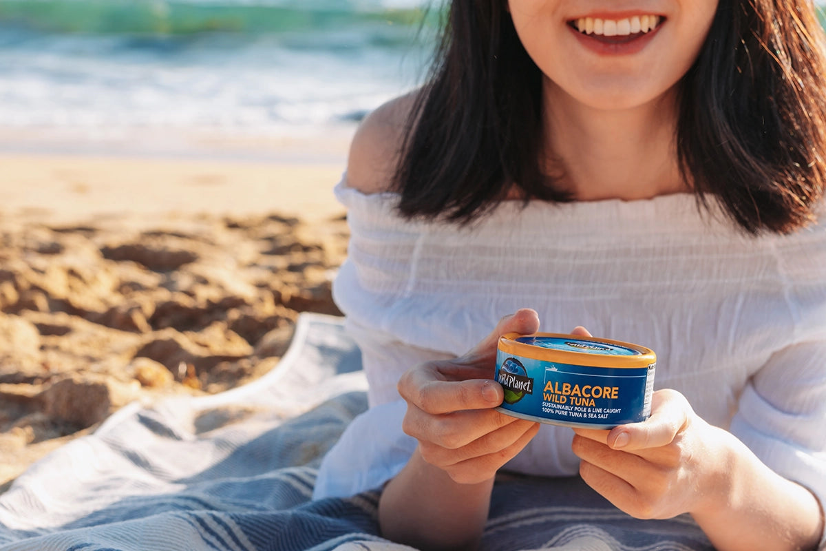 Woman holding a can of Wild Planet Albacore Wild Tuna on a beach in front of ocean
