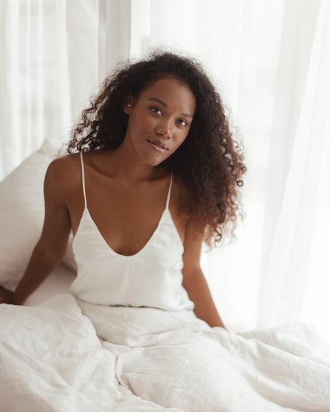 Image is of model Zoya sitting on soft white bedding and looking serenely up at the camera. She is wearing the Dee ivory silk camisole top.